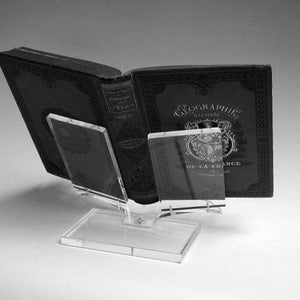 Op de afbeelding: Een open antiek boek met sierlijke omslagdetails, ondersteund door een heldere acryl boekenstandaard. Het boek heet "GEOGRAPHIE" en "DE LA FRANCE". De standaard heeft een rechthoekige basis en schuine steunen.