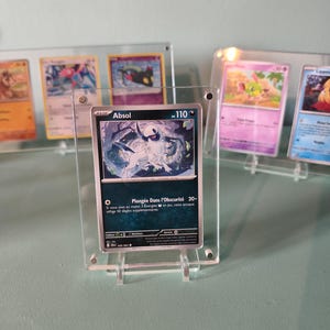 Peut inclure: Une carte de jeu Pokémon avec Absol, présentée dans un support en acrylique transparent. La carte présente une illustration en noir et blanc d'Absol avec le texte "Absol" et "110". D'autres cartes Pokémon sont visibles en arrière-plan.