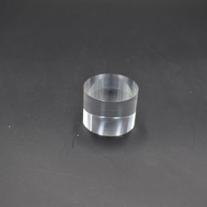 May include: A clear, cylindrical object made of transparent material. The object is a short cylinder with a flat top and bottom. The item is sitting on a dark surface. The object is likely a display stand or a decorative element.