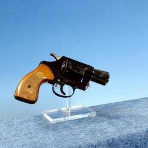 Op de afbeelding: Een zwarte revolver met een houten handvat wordt getoond tegen een blauwe achtergrond. Het pistool is gemonteerd op een heldere acryl standaard. De handgreep heeft een lichtbruine, getextureerde uitstraling.