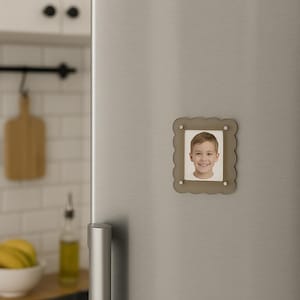 May include: A small, scalloped-edged, gray photo frame with a picture of a smiling child attached to a refrigerator. The frame is held in place by four small, silver-colored magnets.