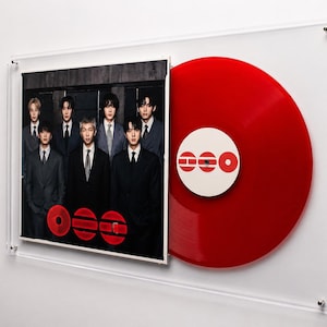 Peut inclure: Un cadre mural en acrylique transparent contenant une pochette d'album carrée montrant sept personnes en costume sombre et un disque vinyle rouge vif. Le disque possède une étiquette centrale blanche avec le texte rouge "BTS" en police grasse.
