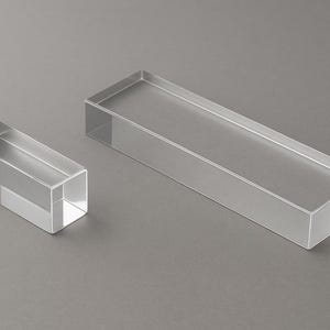 Peut inclure: Deux blocs d'exposition rectangulaires en acrylique transparent. L'un est plus petit, d'environ 7,6 cm de long, et l'autre est plus grand, d'environ 22,9 cm de long. Les deux ont des dessus ouverts et sont posés sur un fond gris.