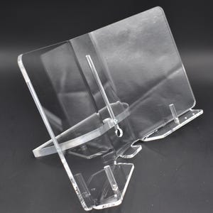 Peut inclure: Support de livre en acrylique transparent avec une base rectangulaire et un support incliné. Le support présente un design clair, adapté à l'affichage de livres ou d'autres articles. Le support est conçu pour maintenir les articles en position verticale.