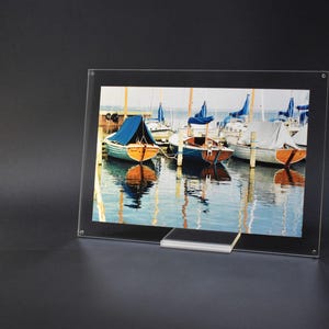 Peut inclure: Un cadre photo en acrylique transparent avec une image de bateaux amarrés dans un port. Les bateaux ont des voiles bleues et blanches et se reflètent dans l'eau. Le cadre a un support transparent.