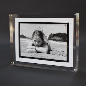 Peut inclure: Cadre photo en acrylique transparent avec une photographie en noir et blanc d'un enfant allongé sur une plage. Le cadre a un bord blanc et est maintenu par de petites vis argentées. Le fond est gris foncé.