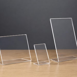Pode incluir: Três suportes para sinalização em acrílico transparente de vários tamanhos e orientações. Um suporte está posicionado horizontalmente, enquanto os outros dois estão verticais, com um design inclinado e base plana para estabilidade.
