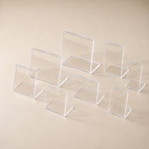 Pode incluir: Uma coleção de oito suportes para sinalização em acrílico transparente em vários tamanhos retangulares. Cada suporte apresenta um design em forma de L com uma base plana e um painel vertical para exibir placas, menus ou fotografias.