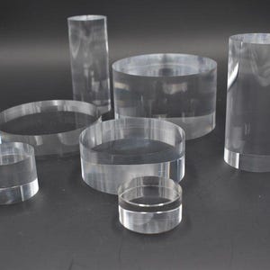 Op de afbeelding: Een verzameling heldere acryl cilinders en schijven van verschillende hoogtes en diameters. De objecten zijn gerangschikt op een donkere ondergrond, wat hun transparantie en gladde oppervlak benadrukt. De cilinders variëren van korte, brede vormen tot hoge, slanke.