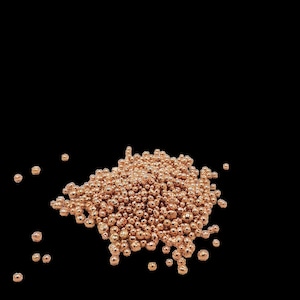 Pode incluir: Um monte de pequenas contas redondas de cor cobre. As contas estão espalhadas sobre um fundo preto.
