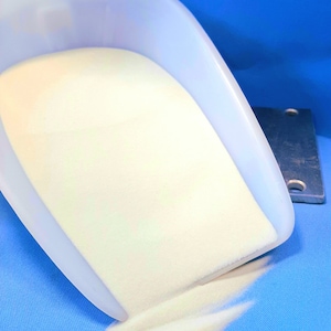 Könnte beinhalten: Eine weiße Plastikschaufel gefüllt mit weißem Pulver, das auf eine blaue Oberfläche rieselt.