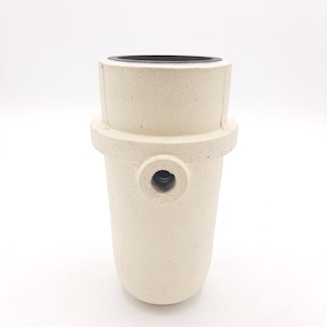 May include: A cylindrical, cream-coloured ceramic object with a black interior rim and a small hole on the side. The object has a textured surface and a rounded base, set against a white background.