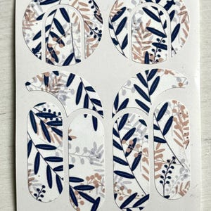 以下が含まれることがあります： ボタニカルデザインのネイルアートステッカーシート。ステッカーは、白の背景にネイビーブルー、ライトブラウン、グレーの葉と花のパターンがあります。ステッカーは、アーチや長方形など、さまざまな形をしています。
