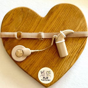May include: A heart-shaped wooden plaque with a beige strap and a beige cochlear implant device. The device includes a round component and a cylindrical component. A small sticker with two cartoon girls and the text "A&R twinsies" is on the bottom.
