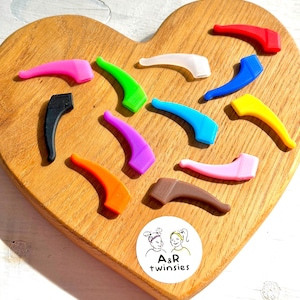 Peut inclure: Un ensemble de douze embouts de branches de lunettes en silicone de différentes couleurs, dont rouge, bleu, jaune, orange, rose, violet, vert, noir, marron et blanc translucide. Un autocollant circulaire présente un dessin au trait et le texte A&R twinsies.