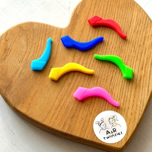 Puede incluir: Una colección de coloridas puntas de patillas de gafas de silicona curvadas en rojo, azul, verde, amarillo y rosa. Las puntas están dispuestas sobre una tabla de madera en forma de corazón. Una pegatina con el texto "A&R twinsies" está en la esquina inferior derecha.