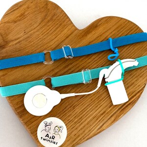 Puede incluir: Dos accesorios para audífonos azules y turquesas se muestran en una tabla de madera en forma de corazón. Los accesorios incluyen correas ajustables con cierres de plástico transparente y un dispositivo blanco con un cable. También es visible una pequeña pegatina con el texto "A&R twinsies".