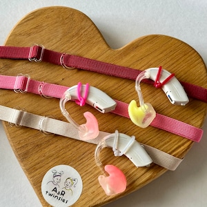Può includere: Tre apparecchi acustici bianchi con diversi colori di auricolari, rosa, giallo e arancione, sono esposti su una superficie di legno a forma di cuore. Gli apparecchi acustici sono attaccati a elastici colorati.