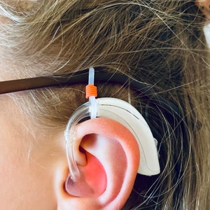 May include: A white behind-the-ear hearing aid with a clear plastic hook and an orange rubber band. The hearing aid is worn on the right ear of a person with blonde hair.