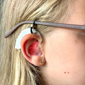 May include: A close-up of a person's ear with a hearing aid attached to a pair of glasses. The hearing aid is white and clear, and the glasses are brown.