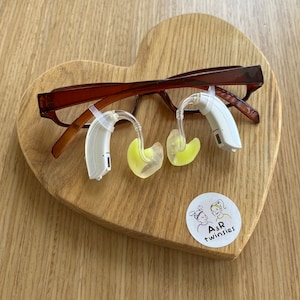 Puede incluir: Dos audífonos blancos con puntas de oído amarillas descansan sobre una superficie de madera marrón en forma de corazón. Un par de gafas marrones descansa sobre los audífonos. Una pegatina blanca con una imagen de dibujos animados de dos niños y el texto "A&R twinsies" está en la superficie.