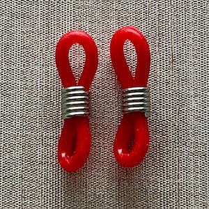 Puede incluir: Dos retenedores de gafas rojos con espirales de metal plateado. Los retenedores están hechos de un material flexible similar al caucho y están diseñados para engancharse alrededor de las patillas de las gafas para mantenerlas seguras. El fondo es una tela gris texturizada.
