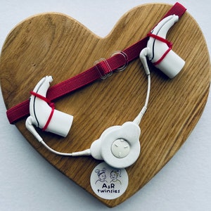 May include: Two white hearing aids with red cords attached to a white transmitter on a heart-shaped wooden board. The board has a sticker with the text "A&R twinsies".