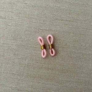 Op de afbeelding: Twee lichtroze siliconen lussen ontworpen voor brillenkoordjes, elk voorzien van een centrale goudkleurige metalen spiraal. Deze kleine, flexibele verbindingsstukken hebben een langwerpige ovale vorm en dienen om brillenpoten vast te zetten.