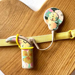 May include: A hearing aid and cochlear implant device with a floral pattern, connected by a white wire, are secured by a yellow elastic band on a wooden heart-shaped surface. The devices feature a mix of pastel colors.