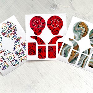 Puede incluir: Tres juegos de pegatinas de arte de uñas en tarjetas blancas. Uno presenta líneas onduladas coloridas, otro diseños de calaveras rojas con detalles en negro, y el tercero patrones de mármol verde, dorado y marrón. Las pegatinas están sobre una superficie de madera blanca.