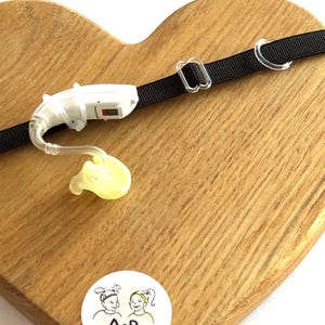 Op de afbeelding: Een wit achter-het-oor hoortoestel met een doorzichtig plastic oorstukje en een dun slangetje, bevestigd aan een zwarte elastische bevestigingsband met een doorzichtige plastic schuifregelaar. Een sticker bevat de tekst A&R twinsies.