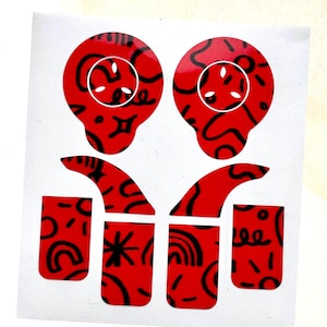 May include: Red stickers with black abstract designs. The stickers feature two skull-shaped designs with white circles and dots, along with various shapes and patterns. The stickers are on a white background.