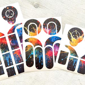 Op de afbeelding: Drie vellen nail art stickers met een zwarte achtergrond en kleurrijke verfspatontwerpen in oranje, geel, blauw en paars. De stickers hebben verschillende vormen, waaronder cirkels, bogen en rechthoekige vormen.