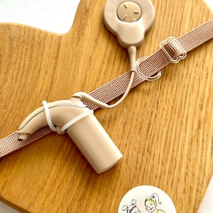 Puede incluir: Un audífono beige con un cable beige y un transmisor beige sobre una superficie de madera en forma de corazón. El transmisor tiene un pequeño botón beige. El cable está unido a una correa beige con una hebilla. La superficie de madera tiene una pegatina con un dibujo de dos chicas y el texto "A&R twinsies".