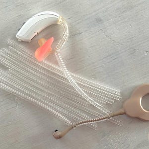 Può includere: Una collezione di accessori per apparecchi acustici, tra cui un apparecchio acustico bianco, inserti auricolari rosa e tubi trasparenti e scanalati. È presente anche un ricevitore beige. Gli articoli sono disposti su una superficie chiara.