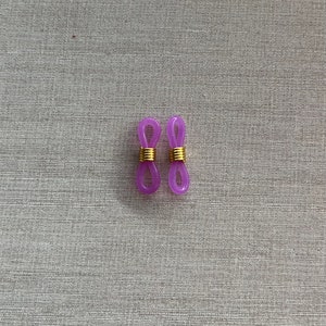 Op de afbeelding: Twee paarse siliconen brilkettingverbinders met een goudkleurige metalen spiraalveer in het midden. Elke verbinder heeft aan beide uiteinden een lus, ontworpen om de pootjes van een bril vast te maken aan een ketting of koord.