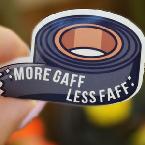 K&ouml;nnte beinhalten: Ein Aufkleber mit einer Rolle dunkelblauem Klebeband mit brauner Mitte. Der Aufkleber tr&auml;gt den wei&szlig;en Text "MORE GAFF LESS FAFF". Der Aufkleber wird in einer Hand gehalten.
