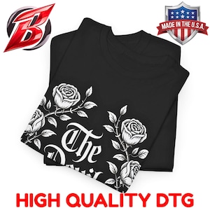 Puede incluir: Camiseta negra con un diseño de rosas blancas y el texto "The Devil". La camiseta está doblada, con el diseño visible en la capa superior. Las palabras "HIGH QUALITY DTG" están impresas en la parte inferior. Un emblema "Made in the U.S.A." está en la esquina superior derecha.