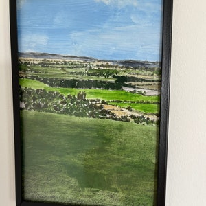 May include: A framed landscape painting in a black metal frame. The artwork shows a green field, trees, and a blue sky with clouds. The painting is suspended on a white wall.