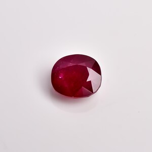 May include: A loose, cushion-cut ruby gemstone with a deep, saturated red colour. The stone features a faceted surface that reflects light, showcasing a translucent quality and a polished finish. The gem has a rounded, rectangular shape.