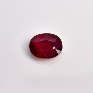May include: A deep red, oval-cut gemstone with a faceted surface. The stone displays a rich, saturated crimson hue and exhibits internal light reflections across its polished, multi-faceted top, creating a clear and brilliant appearance.