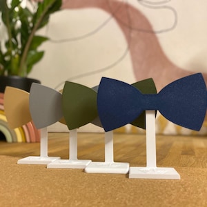 Puede incluir: Cinco pajaritas expuestas en soportes blancos. Las pajaritas son de diferentes colores: beige, gris, verde, azul y azul marino.