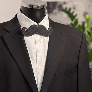 Puede incluir: Una chaqueta de traje negra con una camisa blanca de cuello y una pajarita de fieltro negro. La pajarita tiene forma de bigote.