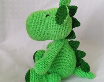 May include: A green crocheted dinosaur toy with a large head and a long tail. The dinosaur has a small, black eye and a green, crocheted spine.