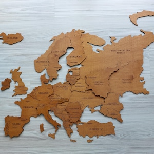 Peut inclure: Une carte murale en bois de l'Europe présentant des formes de pays découpées au laser et des étiquettes gravées pour les nations et les régions. La carte est composée de pièces en bois individuelles assemblées pour former le continent.