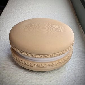 May include: A beige, 3D-printed macaron cookie. The top and bottom halves are a light tan color with a textured, concentric circle design. The filling is white. The edges of the cookie have a decorative, ridged pattern.