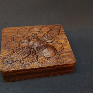 Puede incluir: Una caja de madera con un diseño tallado de abeja y panal. La abeja está en el centro de la caja y mira hacia la derecha. El panal rodea la abeja.