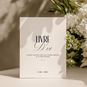 以下が含まれることがあります： 黒い筆記体で「LIVRE D'or」と書かれた白い本。テキスト「LAISSEZ UN PETIT MOT AUX JEUNES MARIES, M. ET MME DUPONT」が下にあります。背景には白い花があります。