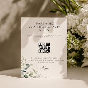 May include: A white card with French text, including "PARTAGEZ VOS PHOTOS AVEC NOUS!" and a QR code. The card provides instructions to download the Wedshoots app and enter an album code. White flowers and foliage are in the background.