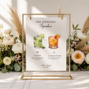 Può includere: Una cornice dorata in metallo contiene un cartello stampato con illustrazioni ad acquerello di due cocktail: un Mojito e un Boulevardier. Il cartello recita "Nos Cocktails Signature" e include i nomi e gli ingredienti dei cocktail. Composizioni floreali circondano la cornice.